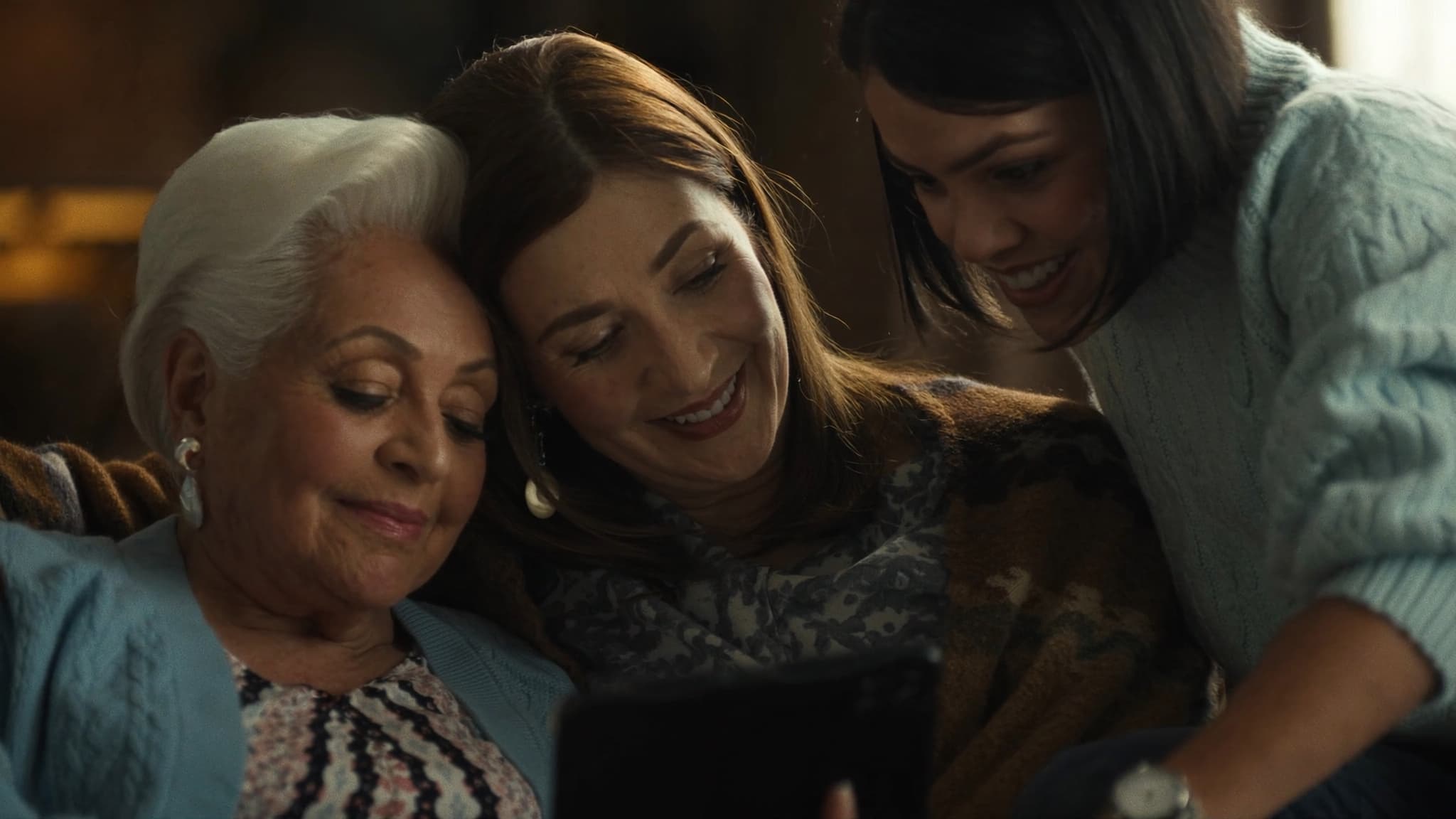 Tres generaciones de familia latina compartiendo una tablet en la sala con luz cálida